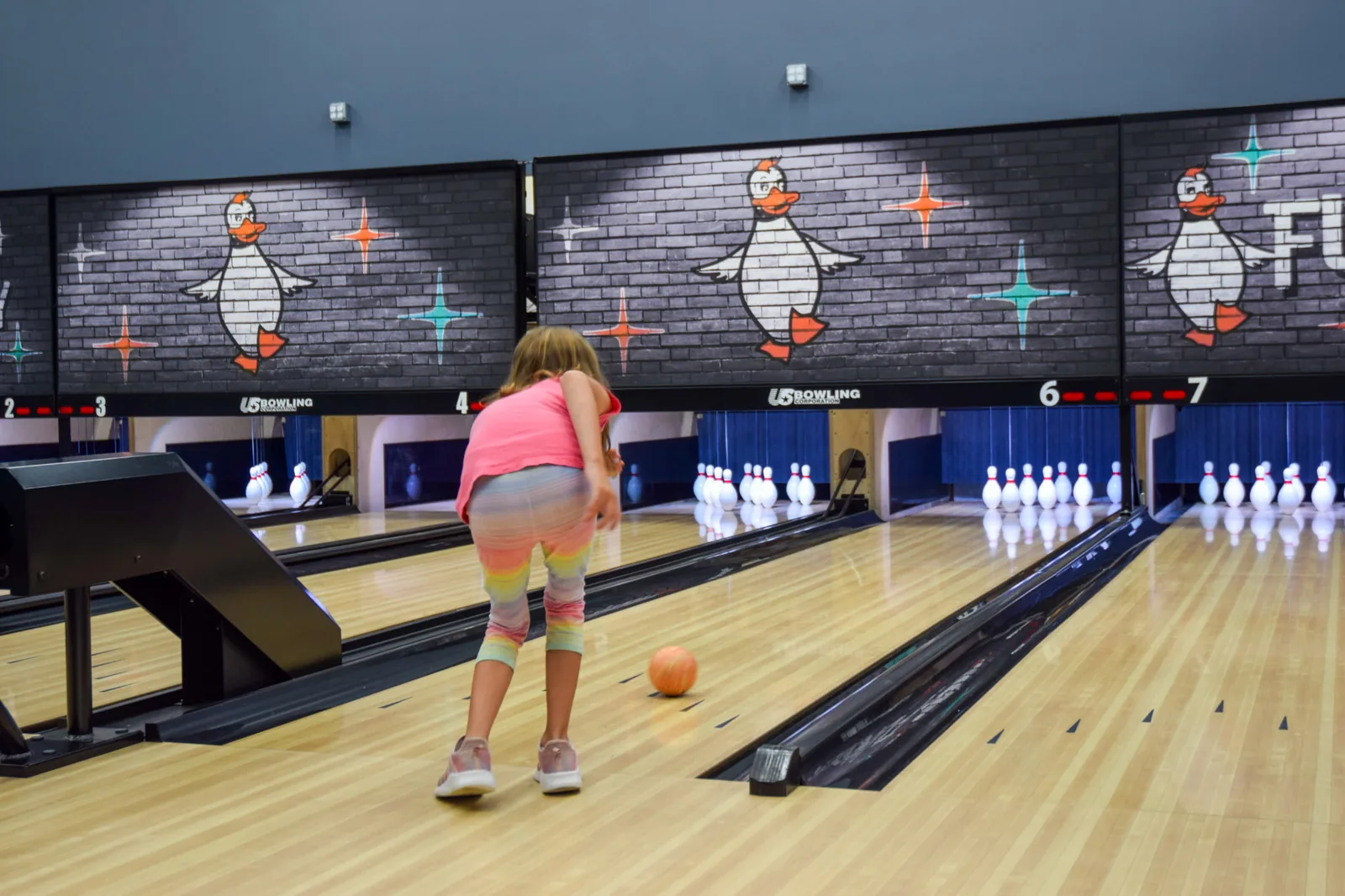 A young bowler rolling a duckpin ball down a Funder Alley lane toward a full rack of pins